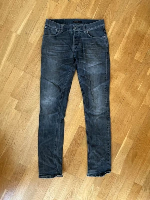 Mörkgrå Grim Tim från Nudie Jeans - Snygga mörkgrå Grim Tim jeans från Nudie Jeans med klassisk femficksdesign och subtila slitningar. Jeansen har normal midja och raka ben, perfekta för en avslappnad men stilren look. Storlek 32/36.
