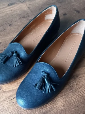 Mörkblå loafers från Alberville med tofsar storlek 35 - Snygga mörkblå loafers från Albertville i läder med klassiska tofsar framtill. Skorna har en rundad tå och en stilren design som passar perfekt till jeans eller kostymbyxor. Fodret är i ljusbrunt läder för extra komfort.