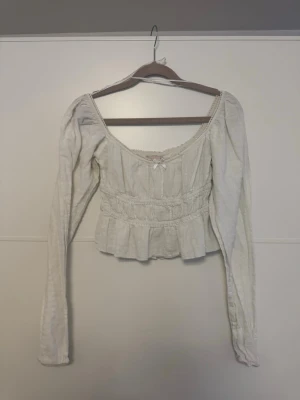 Vit romantisk blus från Nelly - Supersöt vit blus från Nelly med lång ärm, smockad midja och volang nedtill. Blusen har en djup rundad urringning med spetsdetaljer och en liten rosett framtill. Tillverkad i ett lätt, krinklat tyg som ger en romantisk vibe.❤️