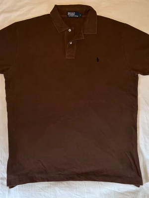 Ralph Lauren | Piké | L - Snygg brun pikétröja från Ralph Lauren med klassisk krage och två knappar framtill. Tröjan har den ikoniska broderade loggan på bröstet och är gjord i ett mjukt bomullsmaterial. Perfekt för en avslappnad men stilren look. Mått: Längd 73 Bredd 58
