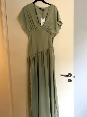 Ljusgrön långklänning från Gina Tricot - Snygg ljusgrön långklänning från Gina Tricot med v-ringning och korta ärmar. Klänningen har en lös passform och faller fint ner till golvet. Perfekt för sommarens tillfällen och enkel att styla med accessoarer.