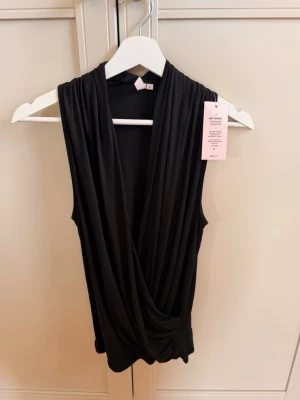 Svart omlottopp från Nelly - Snygg svart omlottopp från Nelly med djup v-ringning och ärmlös design. Toppen har ett flowy fall och är perfekt för att skapa en trendig look. Tillverkad i mjukt och följsamt material som känns skönt mot huden. Helt ny och oanvänd