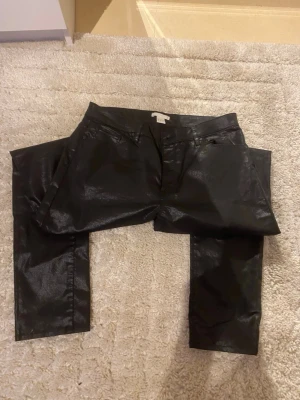 Svarta coated jeans från H&M - Snygga svarta coated jeans från H&M i storlek 42. Byxorna har en glansig finish som ger en cool look och klassisk femficksmodell. Perfekta för dig som vill ha ett par trendiga jeans med lite edge.