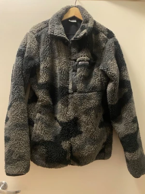 Columbia fleece i camomönster - Fet fleece från Columbia i grått och svart camomönster. Jackan har hög krage, halv dragkedja och en bröstficka med logga. Materialet är supermjukt och fluffigt, perfekt när du vill hålla dig varm. Långärmad och riktigt chill stil.