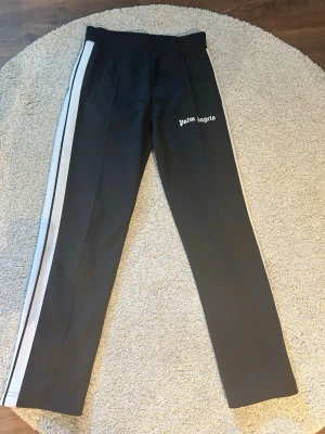 Palm Angels svarta mjukisbyxor - Palm Angels svarta mjukisbyxor med vita sidoränder och logga på vänster lår. Byxorna har rak passform, resår i midjan och är tillverkade i ett mjukt, sportigt material. Perfekta för en avslappnad och trendig streetwear-look.