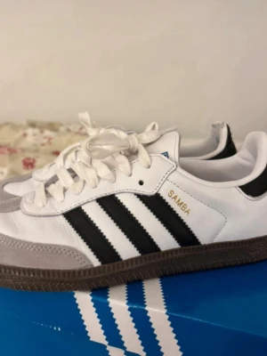 Adidas Samba vita och svarta sneakers - Klassiska Adidas Samba sneakers i vitt läder med svarta ränder och grå mockadetaljer vid tån. Skorna har bruna gummisulor och vit plös med blå Adidas-logga. Perfekta för en sportig och stilren look.