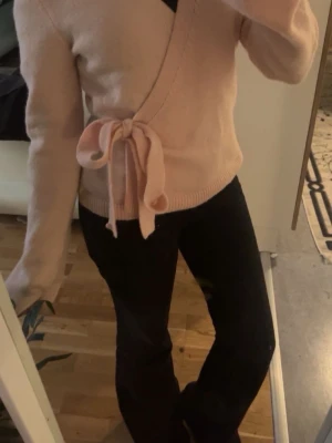 Ljusrosa omlottstickad tröja - Supersöt ljusrosa stickad omlott-tröja med knytband i midjan. Tröjan har långa ärmar och ribbade muddar. Perfekt för lager på lager och ger en feminin touch till din outfit. Tröjan är använd en gång 