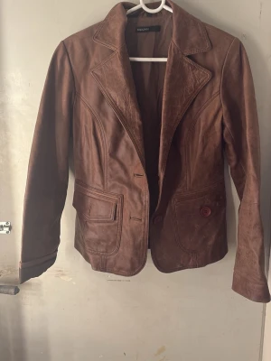 Brun skinnjacka  - Snygg brun skinnjacka med klassisk kavajkrage och två stora fickor framtill. Jackan har två knappar och markerade sömmar som ger en cool vintagekänsla. Perfekt för dig som vill ha en unik och stilren look.