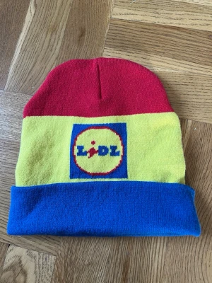 Lidl mössa i retrostil - Säljer en ikonisk mössa i rött, gult och blått med Lidl-loggan framtill. Mössan är stickad och har en uppvikt kant i blått. Materialet är mjukt. Unik mössa då den inte går att köpa längre i butik.