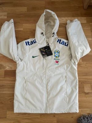 Vit Brasil Nike vindjacka med huva - Snygg vit vindjacka från Nike med Brasilien CBF-logga, blåa Itau-tryck på axlarna och Guaraná Antarctica-detalj. Jackan har huva med dragsko, hel dragkedja och två fickor framtill. Tillverkad i GORE-TEX-material för extra andningsförmåga.