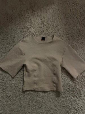Beige croppad topp från Gina Tricot - Trendig beige croppad topp från Gina Tricot med korta ärmar och rund halsringning. Toppens sömmar framtill ger en snygg detalj och den har en figurnära passform. Perfekt att styla med höga jeans eller kjol.