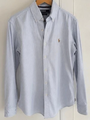 Randig skjorta från Polo Ralph Lauren - Klassisk blå- och vitrandig skjorta från Polo Ralph Lauren med broderad logga på bröstet. Slim fit-modell med lång ärm, knappar framtill och button down-krage. Tillverkad i bomull för en skön känsla.