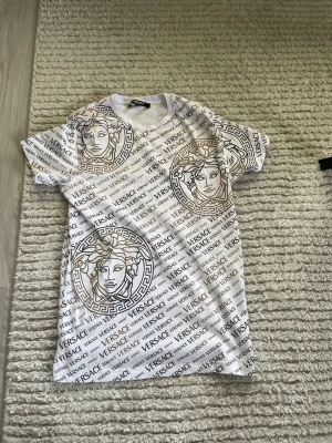 Vit Versace t-shirt med logomönster - Säljer en vit t-shirt från Versace med all-over logomönster i svart och guld. T-shirten har stora grafiska tryck med Medusa-huvuden och klassiska Versace-detaljer. Rund halsringning och korta ärmar. Perfekt för dig som gillar statement-plagg. PRISET KAN DISKUTERAS 
