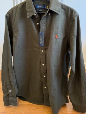 Olivgrön skjorta från Polo Ralph Lauren - Klassisk olivgrön skjorta från Polo Ralph Lauren med orange broderad logga på bröstet. Perfekt för sommaren. Helt ny, aldrig använd. Säljes pga fel strl. Hör av dig vid frågor eller funderingar. Pris kan diskuteras vid snabb affär. 