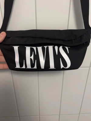 Svart magväska från Levi's - Snygg svart magväska från Levi's med stort vitt logotryck framtill. Väskan har justerbart band och dragkedja upptill. Perfekt för att bära med sig det viktigaste och passar till streetwear-stilen. Aldrig använd.
