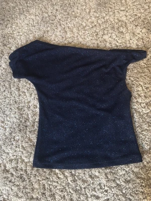 Glittrig mörkblå oneshoulder topp - Snygg mörkblå topp med glittrigt tyg och oneshoulder-design. Perfekt för dig som vill sticka ut med en trendig och unik look. Toppens material är stretchigt och sitter snyggt på kroppen.