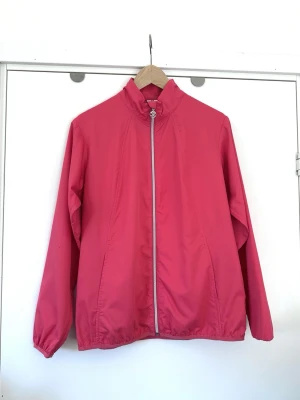 Daily Sports - rosa jacka - storlek L - Stilren rosa träningsjacka / golfjacka / friluftsjacka / jacka från Daily Sports i storlek L. Klassisk modell med dragkedja framtill och en hög krage. Jackan har långa ärmar med elastiska muddar och en elastisk nederkant. Det finns två diskreta fickor i sidorna. Dragkedjan har en dekorativ dragtapp formad som ett hjärta med en golfklubba. Mycket gott skick, som ny!