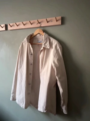 Beige overshirt från Weekday - Stilren beige overshirt från Weekday med klassisk krage och knäppning framtill. Overshirten har långa ärmar och är tillverkad i ett kraftigare bomullstyg som ger en avslappnad och trendig look.