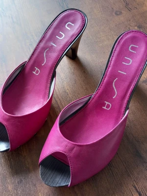 Unisa Rosa Mules i storlek 36 - Ett par rosa mules med öppen tå och klack. Skorna har dekorativa nitar längs sidorna och en slät innersula med Unisa-logotypen.