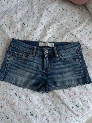 Blå jeansshorts från American Eagle - Snygga blå jeansshorts från abercrombie and fitch med klassisk femficksdesign, låg midja och råa kanter. Shortsen har slitna detaljer och ljus tvätt, perfekt för en avslappnad stil. Passar dig som gillar en casual och trendig look.