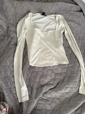Vit långärmad topp med fyrkantig ringning - Snygg vit långärmad topp med trendig fyrkantig ringning. Toppens passform är slim och den är tillverkad i ett mjukt, stretchigt material som känns skönt mot huden. Perfekt att styla med jeans eller kjol för en clean look.