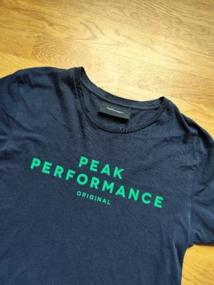 Blå Peak Performance t-shirt - Snygg mörkblå t-shirt från Peak Performance med grönt tryck framtill. Klassisk rund hals och korta ärmar. Tillverkad i mjuk bomull som känns skön mot huden. Perfekt för dig som gillar enkel och stilren design.