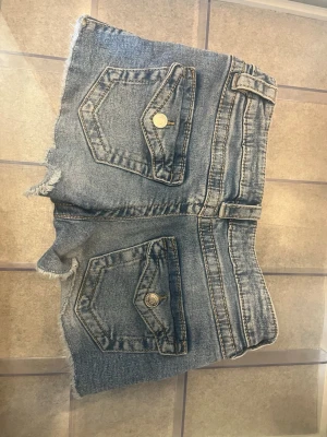 Blå lågmidjade jeansshorts - Säljer ett par blå denimshorts med låg midja och råa, fransiga benslut. Shortsen har två stora fickor bak med knappdetaljer och bälteshällor. Perfekta för sommardagar och en avslappnad stil.