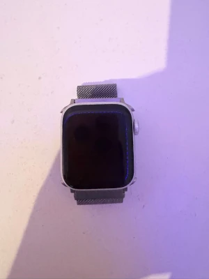 Apple Watch med silver mesh-armband - Snygg Apple Watch med fyrkantig skärm och silverfärgat mesh-armband i metall. Klockan har en digital krona på sidan och laddas trådlöst. Perfekt för dig som vill ha koll på notiser, träning och hälsa direkt på handleden.