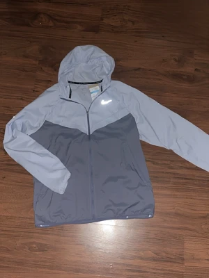 Nike vindjacka  - Säljer en helt ny Nike vindjacka som är oanvänd. Den är i två nyanser av grått med hel dragkedja och huva. Jackan har reflekterande Nike-logga på bröstet och är tillverkad i lätt, vindtätt material. Perfekt för löpning eller utomhusaktiviteter när det blåser.