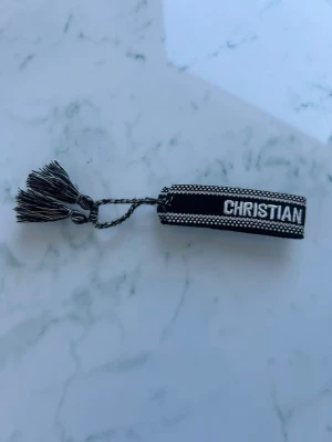 Svart armband Dior - Snyggt svart tygarmband från Dior med texten 'CHRISTIAN DIOR J'ADIOR' i vitt. Armbandet har dekorativa tofsar i ändarna och ett flätat band. Perfekt accessoar för att lyfta din outfit med en exklusiv touch.