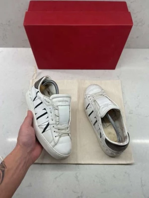Vita Valentino Garavani sneakers - Snygga vita sneakers från Valentino Garavani med svart VLTN-logga på sidorna. Skorna har lågt skaft, vita snören och detaljer i svart på hälen. Tillverkade i läder med klassisk siluett och stilren design.