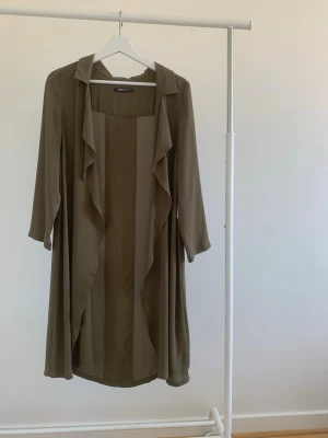 Olivgrön lång kappa med volangkant - Stilren olivgrön kappa från Gina Tricot med långa ärmar och öppen front med volangkant. Lätt och flowigt material som ger en avslappnad look. Perfekt för lager-på-lager och enkel att matcha med olika outfits.