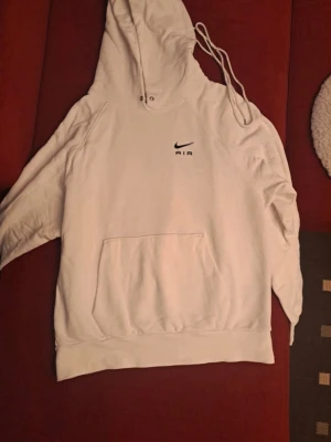 Vit Nike Air hoodie med huva - Säljer en stilren vit hoodie från Nike Air med svart logga på bröstet. Hoodien har en stor känguruficka framtill, dragsko i huvan och ribbade muddar. Perfekt för dig som gillar sportig och avslappnad stil.