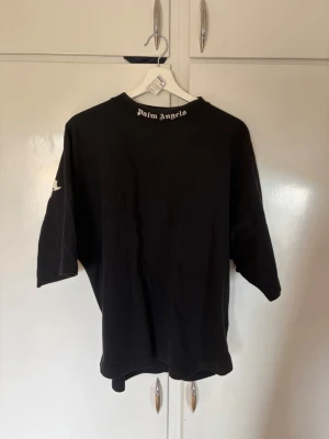 Svart Palm Angels t-shirt - Svart t-shirt från Palm Angels med stort vitt logotryck på ryggen och mindre logga vid halsen. Oversized passform och korta ärmar. Perfekt för dig som gillar streetwear och vill sticka ut med en ikonisk look. Modell på bild är 188 och väger 76kg