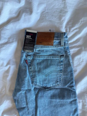 Levi's 501 ljusblå raka jeans - Klassiska Levi's 501 jeans i ljusblå denim med raka ben och knappgylf. Jeansen har fem fickor, läderpatch bak och den ikoniska röda Levi's-taggen på bakfickan. Perfekta för en avslappnad och tidlös stil. Aldrig använda! Lappar kvar och självklart äkta