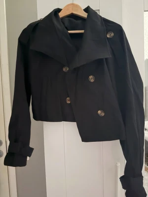 Svart trenchcoat/jacka - Snygg svart trenchcoat/jacka med kortare passform och stora dekorativa knappar framtill. Jackan är helt ny alltså aldrig använd. Jackan har bred krage och uppvikta ärmslut, vilket ger en trendig och modern look. Perfekt att slänga på sig över en hoodie eller t-shirt nu inför sommaren. 