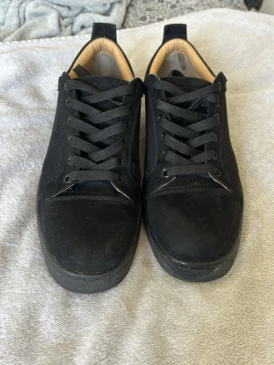 Svarta Louboutin sneakers med röd sula - Snygga svarta sneakers från Christian Louboutin med klassisk röd sula och svarta skosnören. Ovandelen är i mocka och insidan är beige. Perfekta för dig som vill sticka ut med exklusiva detaljer och ikonisk design.