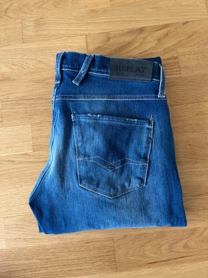 Blå slim fit jeans från Replay - Snygga blå slim fit jeans från Replay med klassisk femficksdesign och diskreta slitningar. Jeansen har en mörkblå tvätt med ljusare partier och detaljerad söm på bakfickan. Perfekt för dig som gillar en modern och stilren look.