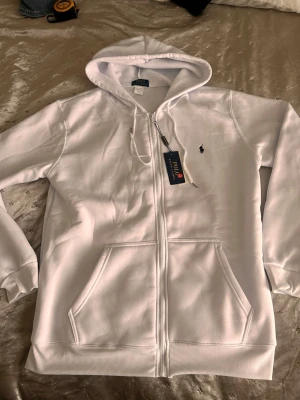 Vit hoodie från Polo Ralph Lauren - Snygg vit hoodie från Polo Ralph Lauren med dragkedja och klassisk logga broderad på bröstet. Hoodien har två stora fickor fram, ribbade muddar och en skön huva med dragsko. Perfekt för dig som gillar stilren design och premiumkänsla.