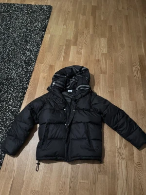 Svart pufferjacka med huva - Säljer en svart pufferjacka med stor huva och dragkedja framtill. Jackan har två stora fickor på sidorna och justerbar dragsko nedtill. Insidan av huvan har ett grått och vitt randigt foder. Perfekt för kalla vinterdagar.