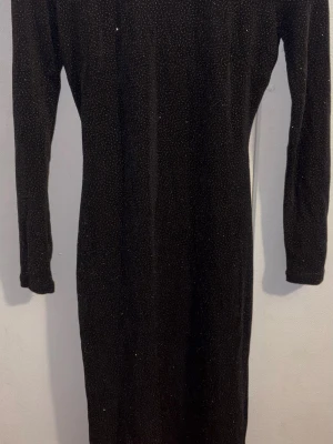 Svart glittrig långärmad klänning - Snygg svart klänning med glitterdetaljer och lång ärm. Klänningen har en tight passform och en cool cut-out med dragkedja i ryggen som ger en unik look. Perfekt för dig som vill sticka ut med stilrena detaljer och trendig design.