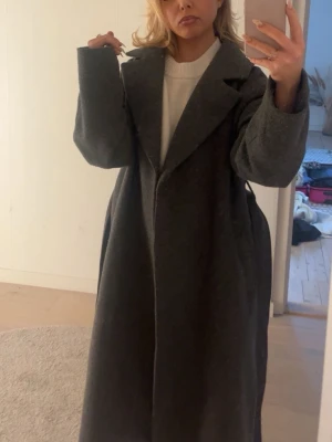 Grå kappa med bälte - Säljer en stilren grå kappa med snörning i midjan. Perfekt för höst och vinter! Den har en krage och långa ärmar, vilket gör den både varm och elegant