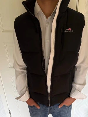 Svart dunväst från Sebago - Snygg svart dunväst från Sebago med hög krage och dragkedja framtill. Västen har en diskret logga på bröstet och en bröstficka med dragkedja. Perfekt att slänga över en skjorta för en clean och stilren look.