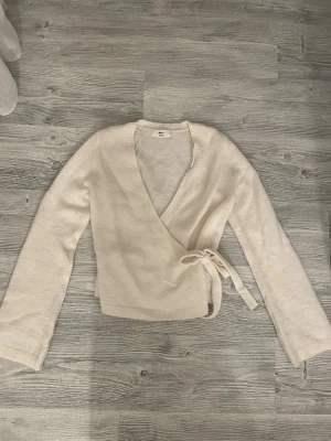 Krämvit omlottstickad tröja - Säljer en krämvit stickad omlottopp med knytband i midjan. Tröjan har långa ärmar, djup v-ringning och ribbade kanter. Perfekt för dig som gillar en enkel men trendig look.