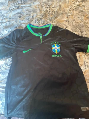 Svart Brasilien fotbollströja Nike - Snygg svart Brasilien fotbollströja från Nike med gröna detaljer och CBF-emblem på bröstet. Tröjan har korta ärmar, rund hals med blå och grön kant samt Dri-FIT-material som andas. Perfekt för träning eller match. Unikt mönster på tröjan
