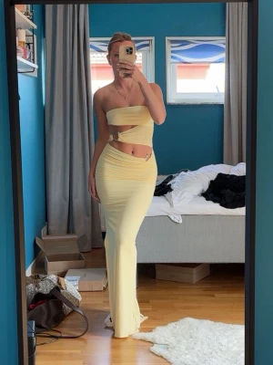 Ljusgul med slits - Trendig långklänning i ljusgul nyans med cutout-detalj och slits. Klänningen är axelbandslös och har en snygg cut-out vid midjan med metallring. Perfekt för dig som vill sticka ut och känna dig extra stylish.