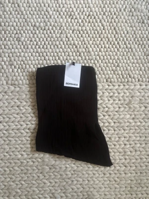 Svart bandeau tubtopp från Bershka - Säljer en helt ny svart bandeau tubtopp från Bershka. Toppen har en ribbad struktur och är axelbandslös, perfekt för en trendig och enkel look. Materialet känns mjukt och följsamt mot huden.