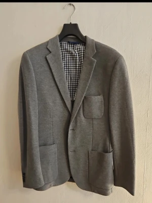 Grå kavaj med rutigt foder - Stilren grå kavaj med tre utanpåliggande fickor och rutigt innerfoder. Klassisk passform med slag och två knappar framtill. Perfekt för en dressad look och enkel att matcha med olika outfits.