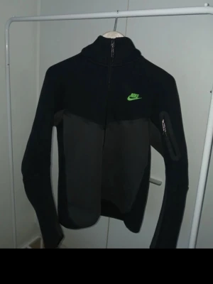 Nike tech fleece - Snygg svart ziptröja från Nike med hög krage och neongrönt Nike-märke på bröstet. Tröjan har dragkedja framtill, en ficka med dragkedja på ärmen och är gjord i ett mjukt, stretchigt material. Perfekt för en sportig och avslappnad stil.
