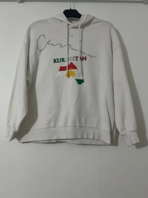 Vit hoodie med Kurdistan-tryck - Vit hoodie med huva och dragsnören. Hoodien har ribbade muddar vid ärmar och nederkant. Perfekt för dig som vill visa ditt stöd för Kurdistan. Priset kan diskuteras. Storleken kan vara stor.